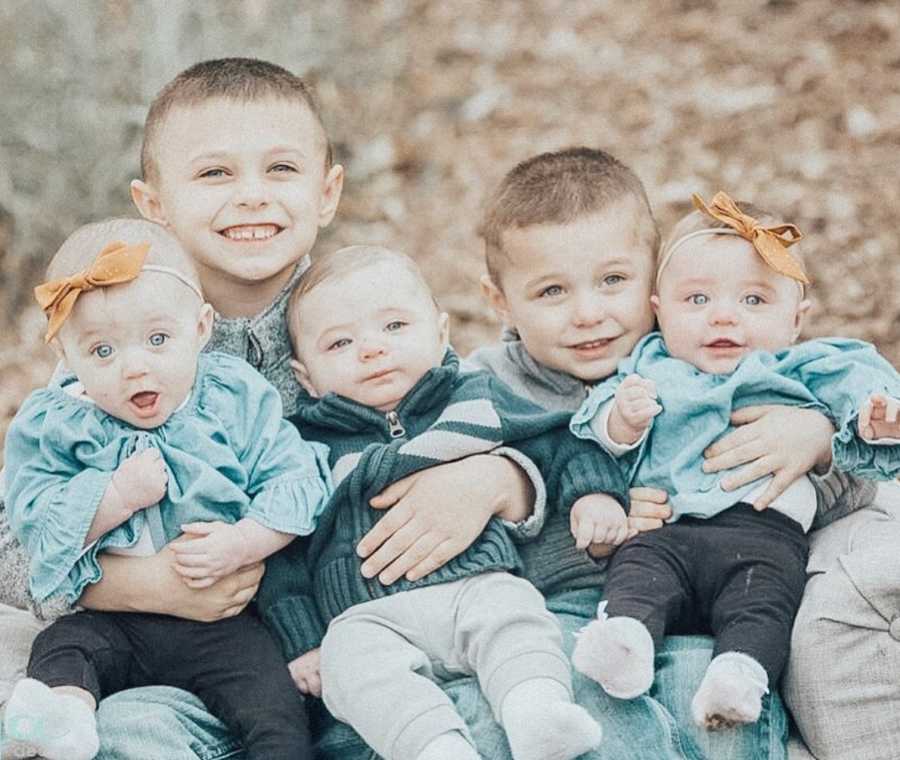 Two young brothers sit on bench outside with their triplet baby siblings in their laps
