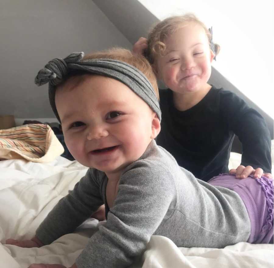 Baby girl lays on her stomach smiling while her older sister with down syndrome sits smiling beside her