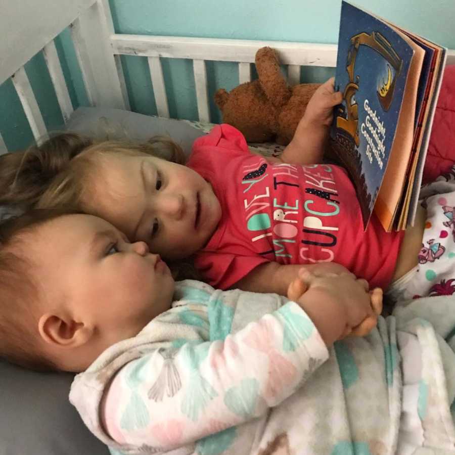 Little girl with down syndrome lays in crib with little sister holding her hand and book