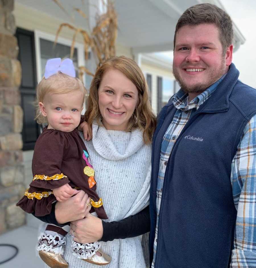 Husband stands beside wife who holds their daughter with mild form of dwarfism outside their house