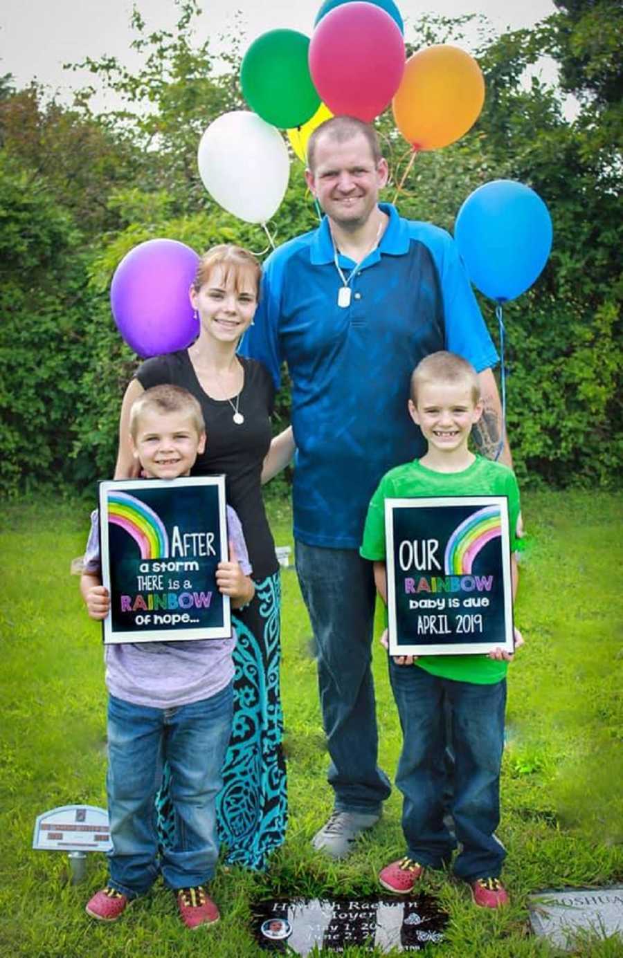 Husband and wife stand in front of balloons with their two sons beside grave of baby that they lost 