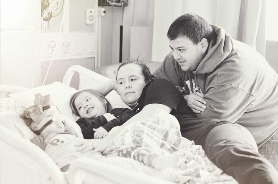 Father leans over hospital bed where his wife and two daughters lay