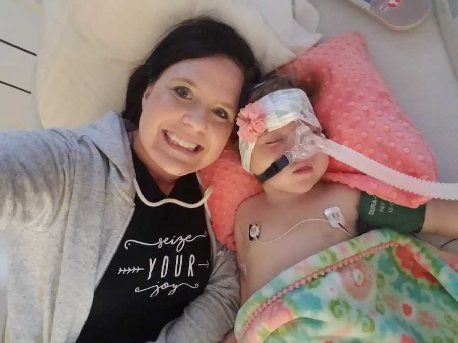 Mother smiles as she takes selfie beside daughter in hospital bed who is hooked up to monitor