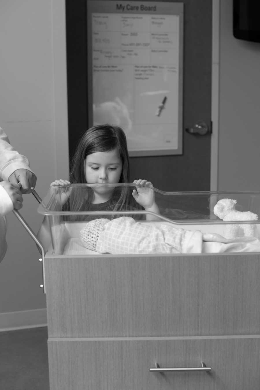 Older sister stands looking over baby sibling who lays asleep in hospital 