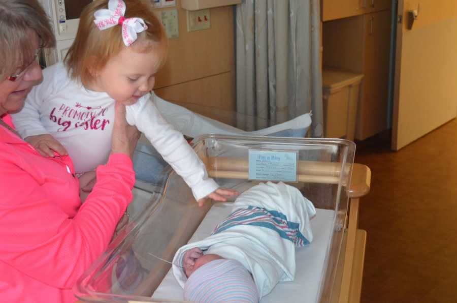 Grandmother holds toddler while they stand above newborn in hospital room