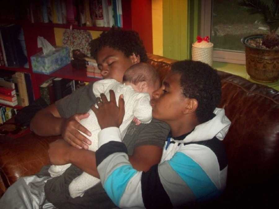 Two teens sit on couch asleep holding baby in their arms