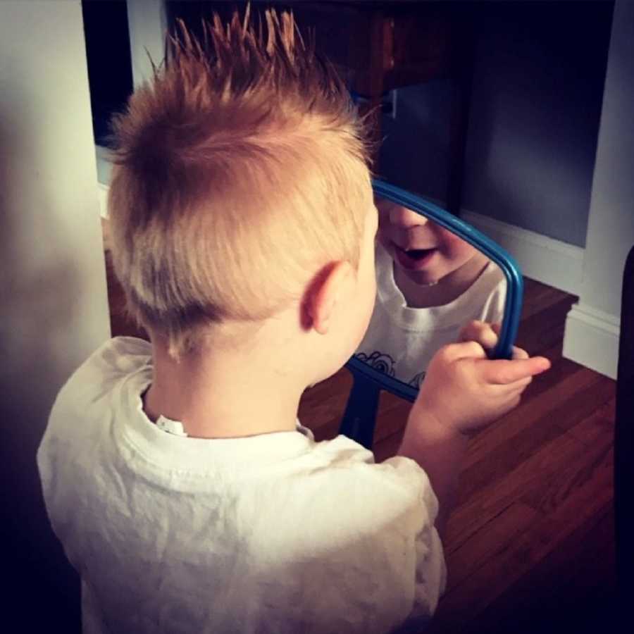Little boy with down syndrome looks into hand held mirror with his hair slicked up in mohawk