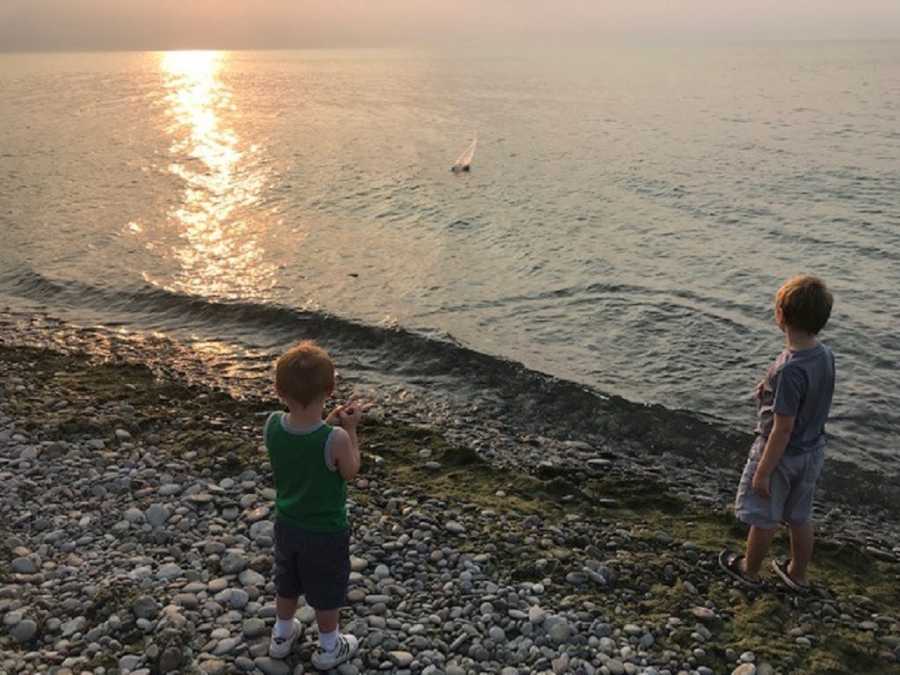 Little boy with down syndrome stands with brother on beach as they watch electric mini sailboat in body of water