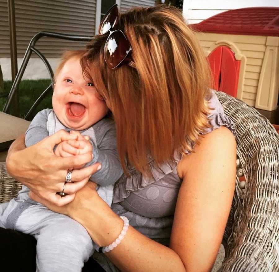 Woman sits in whicker chair outside kissing baby with down syndrome on cheek as he smiles