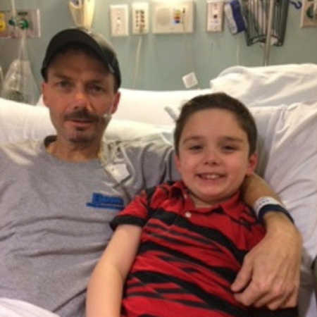 Father with colon cancer sits in hospital bed with arm around his young son