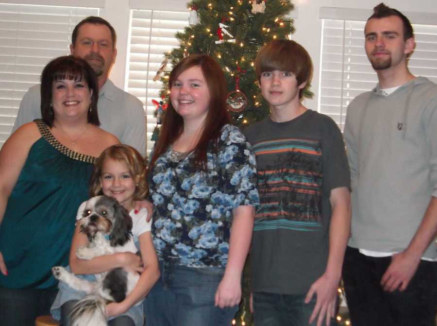 Woman stands in home in front of late husband beside four children, dog and Christmas tree