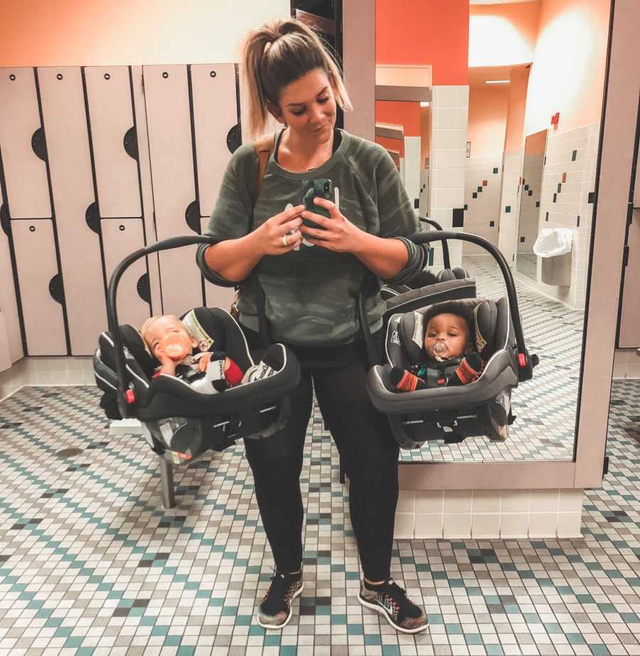 Mother who used to have eating disorder stands in gym locker room taking mirror selfie holding her two kids in car seats