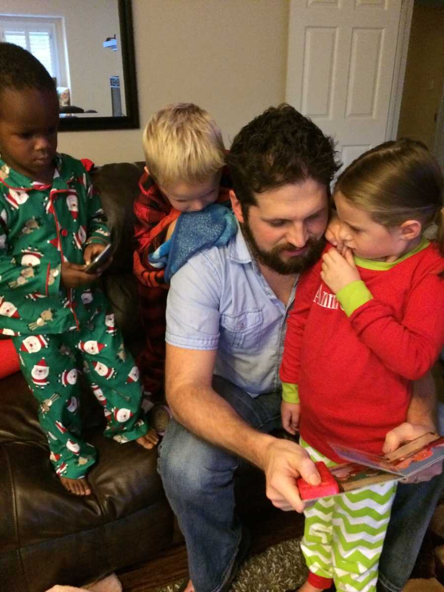 Husband sits on couch reading book to three kids with Christmas pj's on