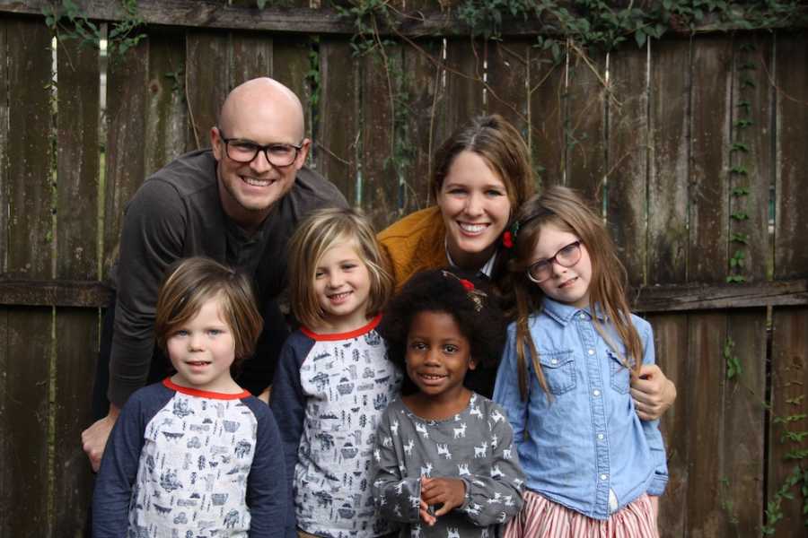 Husband and wife stand outside in front of wood fence with their four children