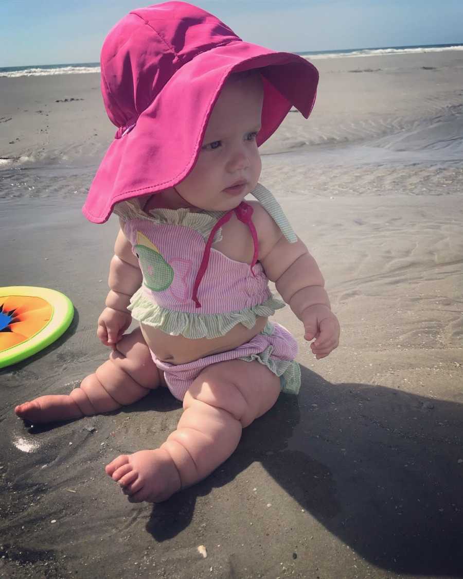 Little girl with mild form of dwarfism sits on beach in bathing suit with hot pink hat on