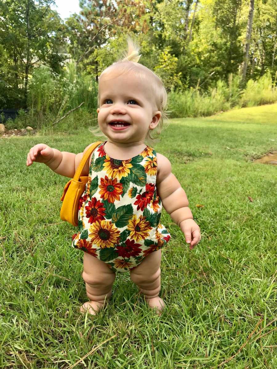 Little girl with mild form of dwarfism stands in yard smiling wearing floral onesie and yellow purse