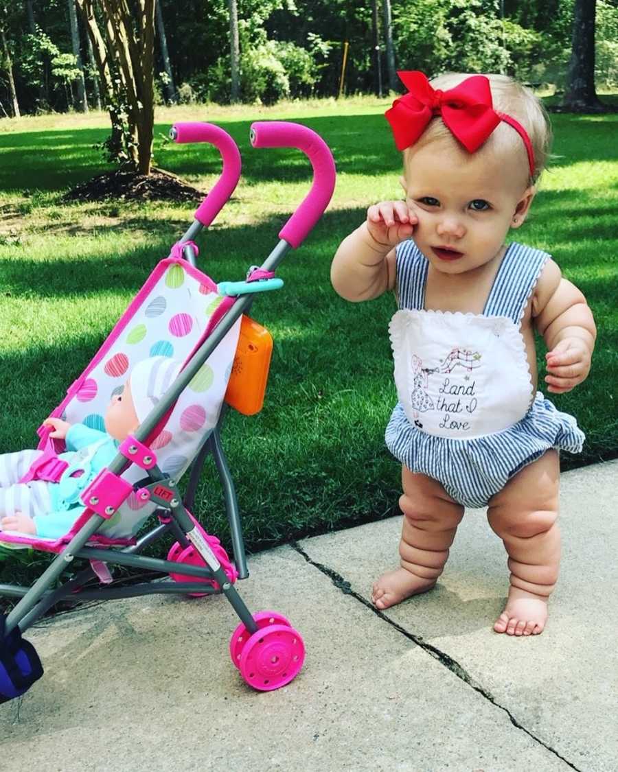 Little girl with mild form of dwarfism stands on sidewalk beside stroller with baby doll in it