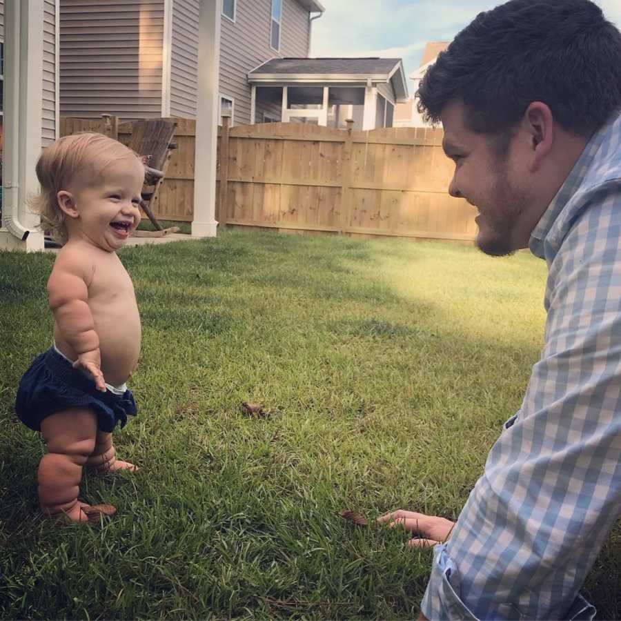 Little girl with mild form of dwarfism stands in yard with only diaper on smiling at dad