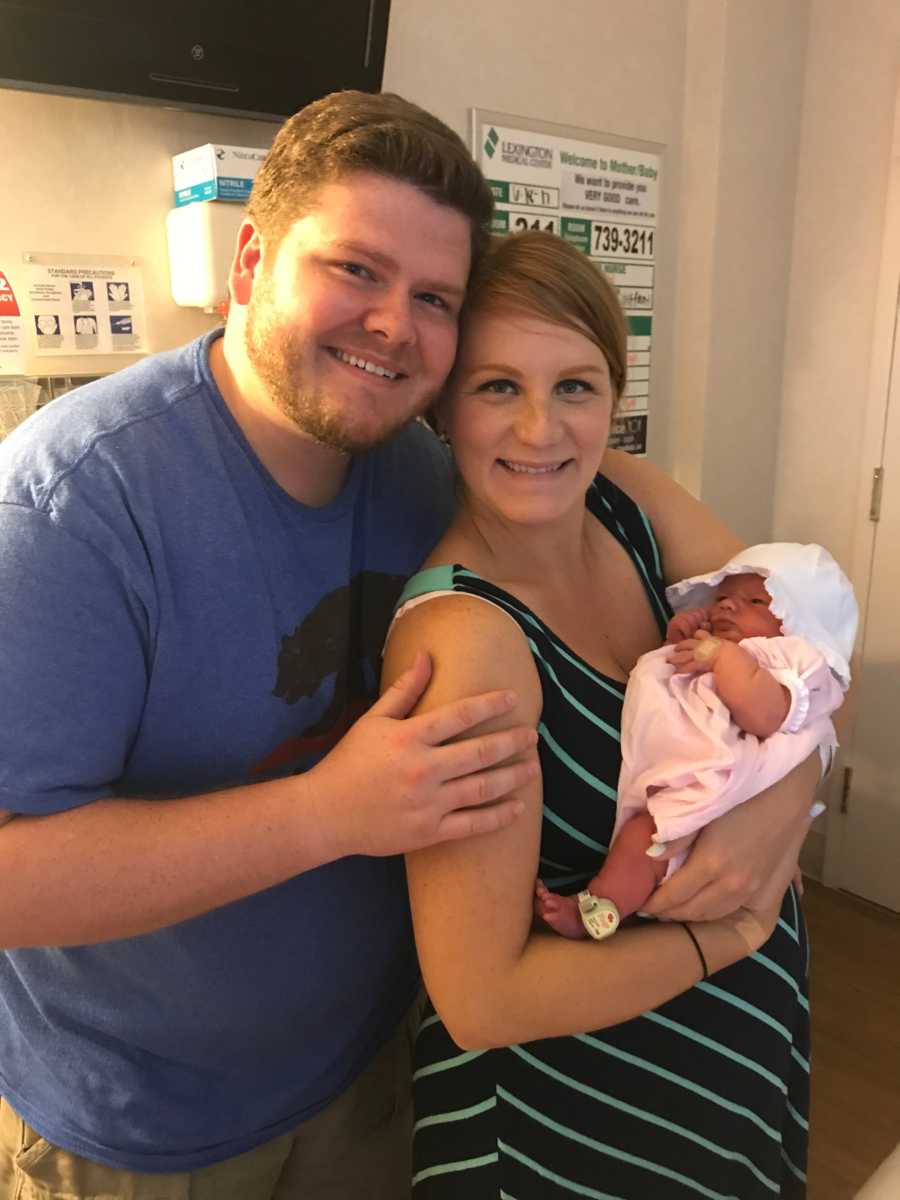 Husband stands smiling beside wife who holds their newborn
