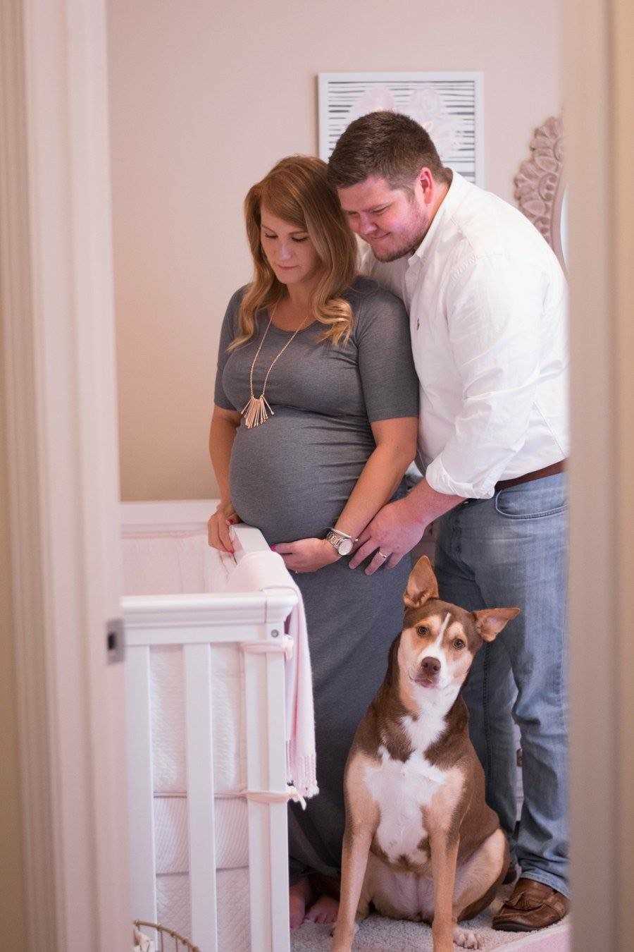 Husband stands behind pregnant wife as they look at crib in baby's nursery beside their dog