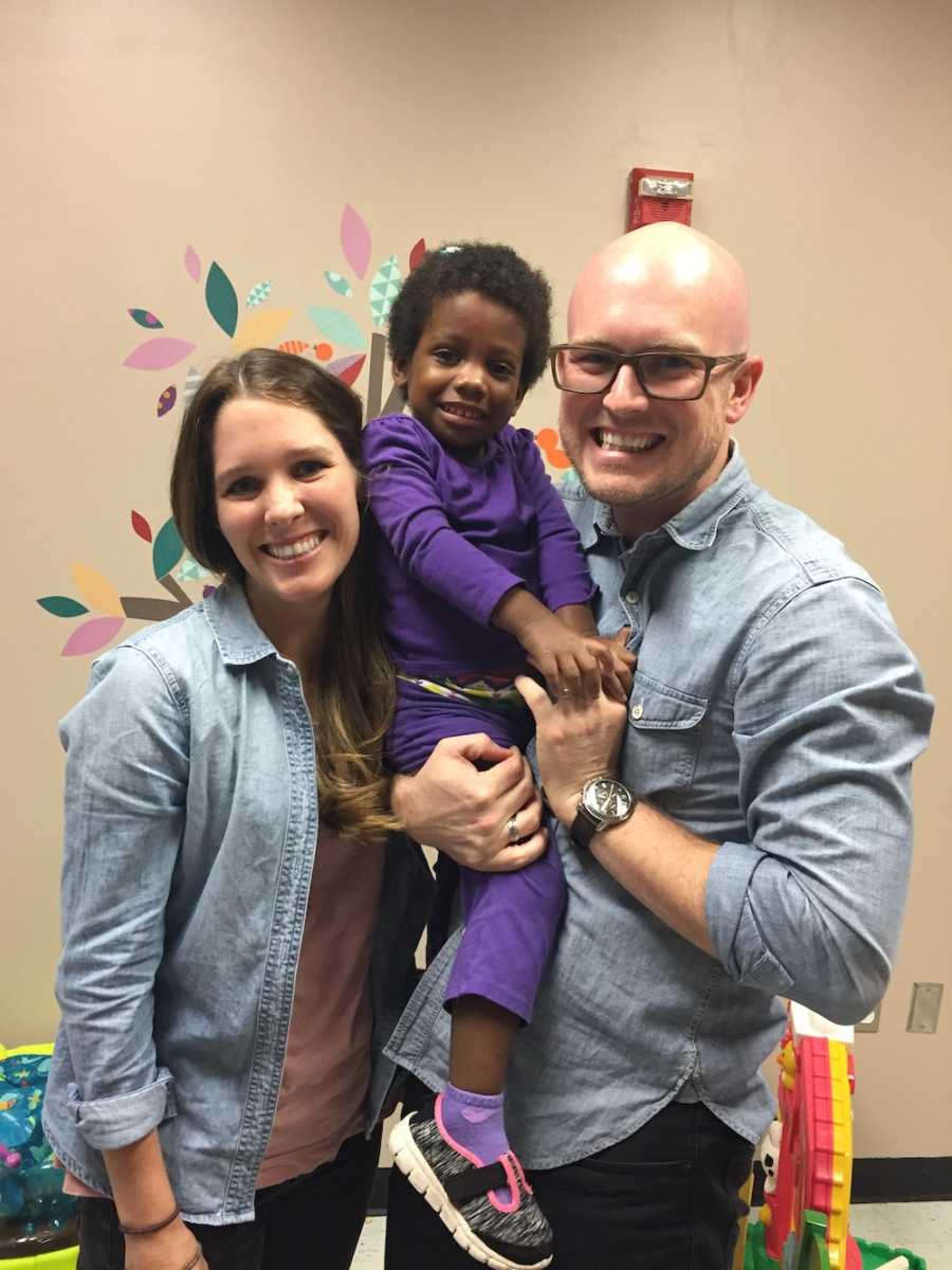 Husband and wife stand smiling while husband holds adopted little girl