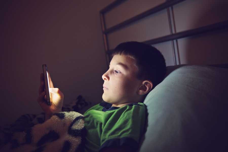 Little boy who is bullied at school lays in bed looking at his phone
