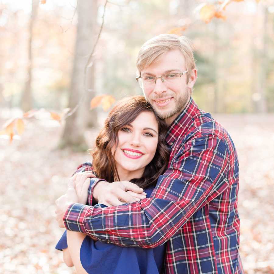 Husband with Acute Disseminated Encephalomyelitis smiles holding wife from behind in woods for photoshoot