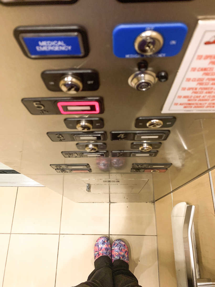 View from above showing woman's legs standing in elevator with floor five button pushed