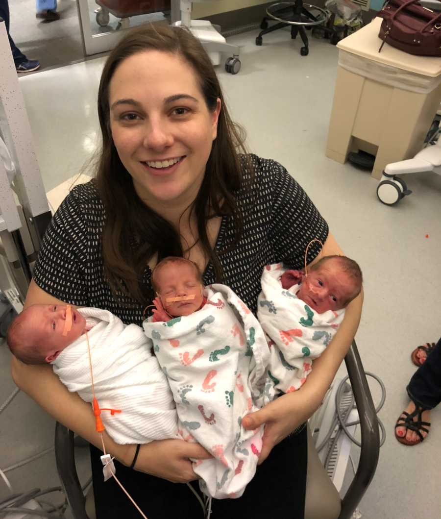 Mother who had preeclampsia sits in chair in NICU smiling as she holds newborn triplets in her arm