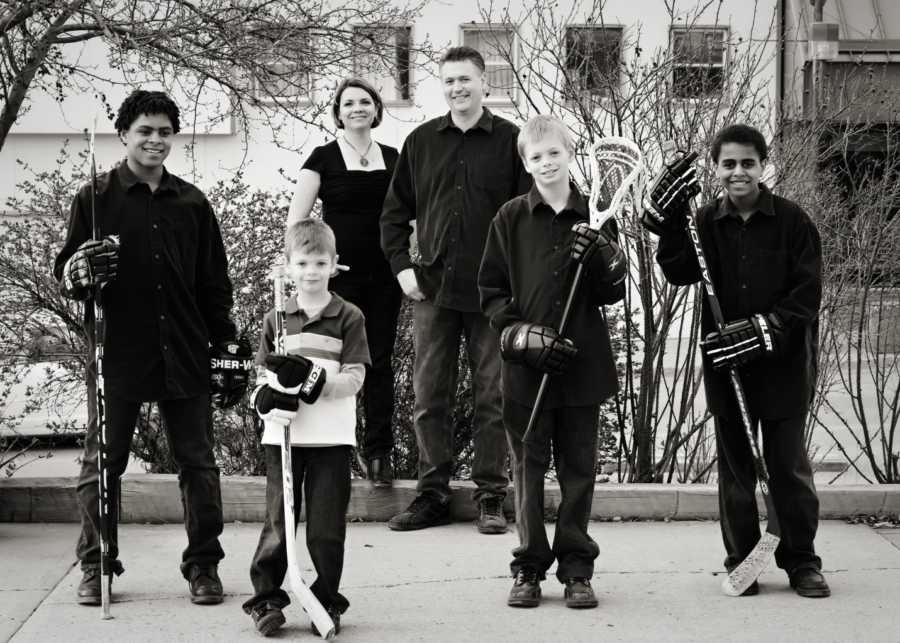 Husband and wife stand on sidewalk beside four sons; three hold hockey sticks and one holds lacrosse stick