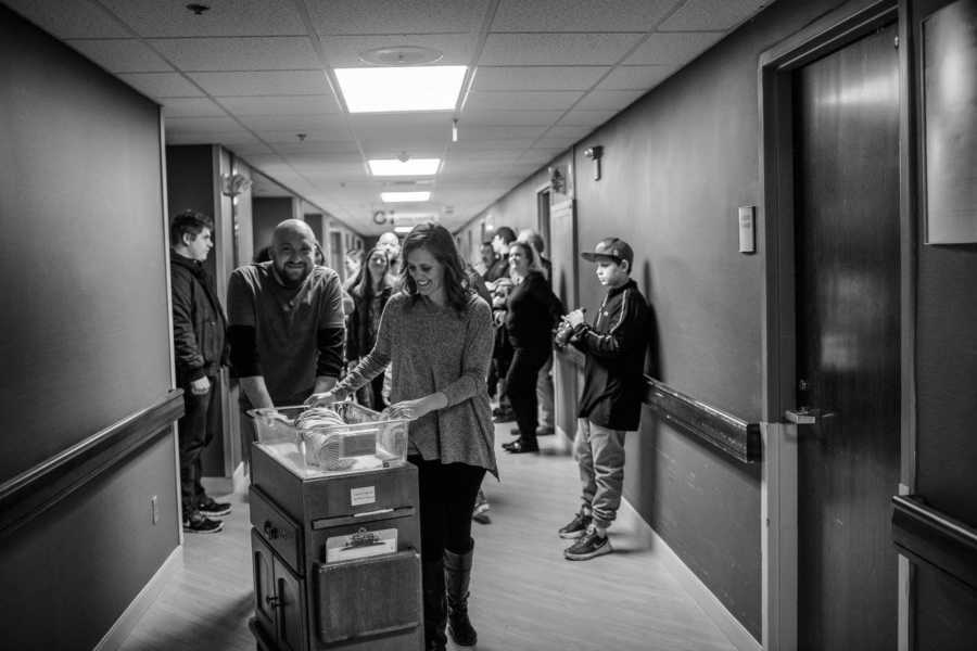 Husband and wife smile as the push adopted newborn in cart down hospital hallway with people behind them