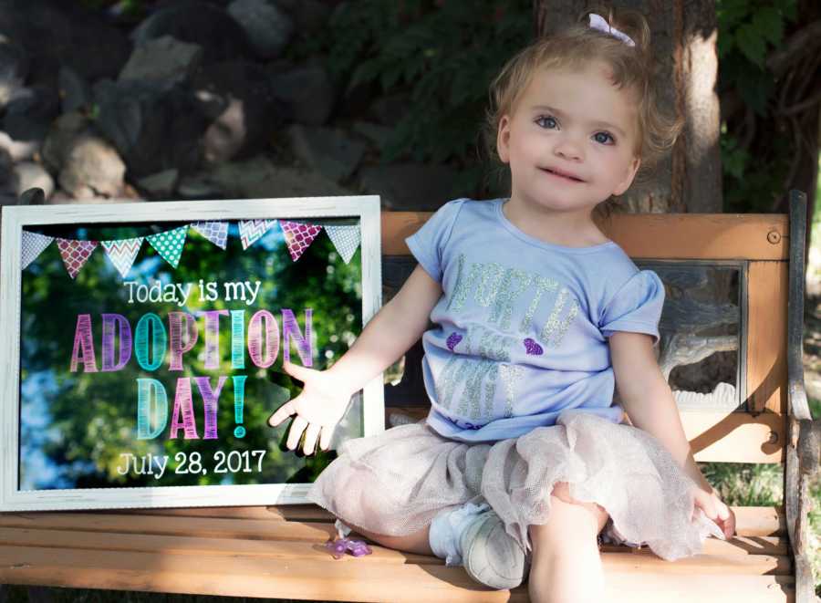 Little girl with Celiac Disease and hyperalgesia sits on bench smiling beside sign that announces her adoption
