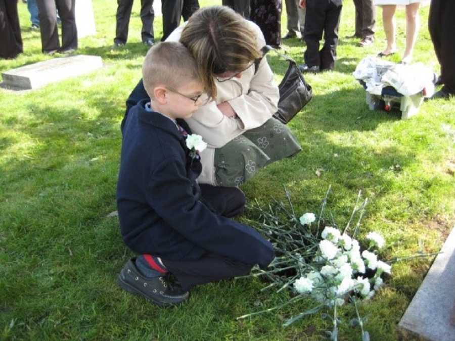 Woman squats beside young son as they look at white flowers at foot of woman's grave