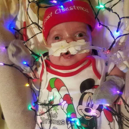 Baby in PICU lays in Christmas hat and onesie surrounded by colorful Christmas lights