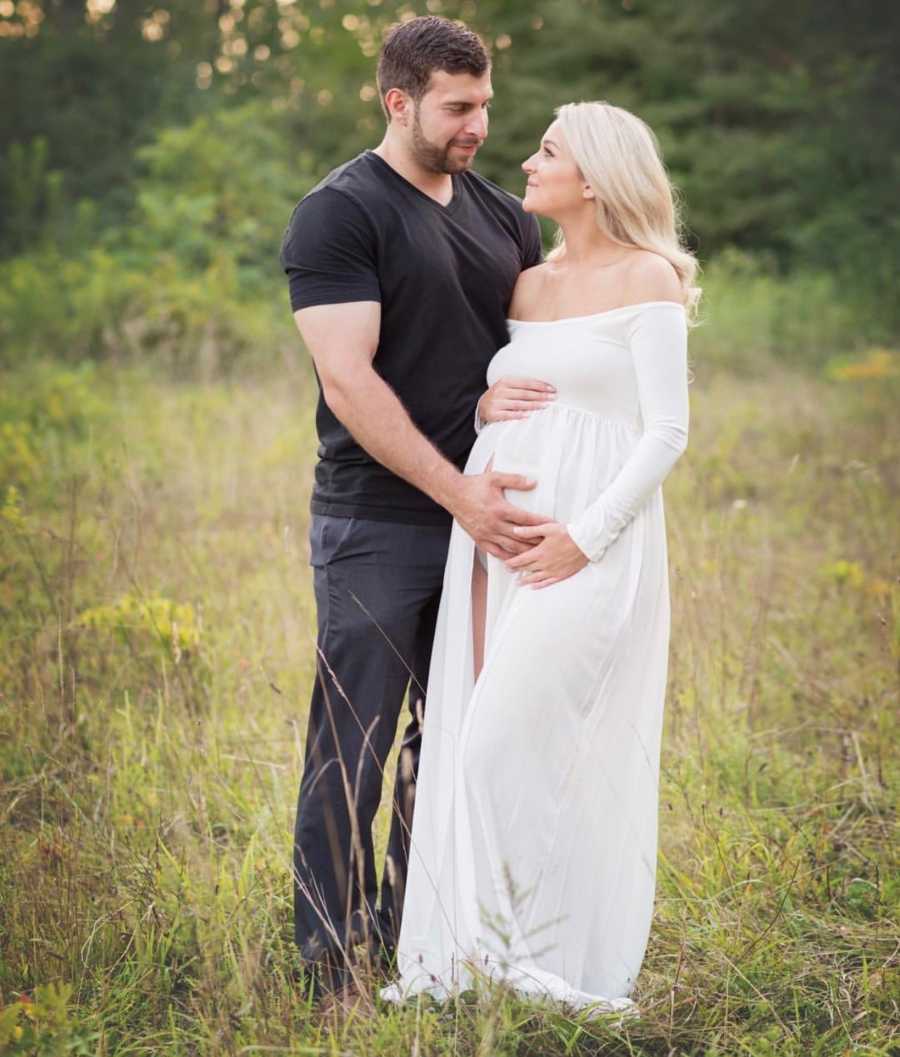 Pregnant woman stands in field holding her stomach with husband who stands beside her