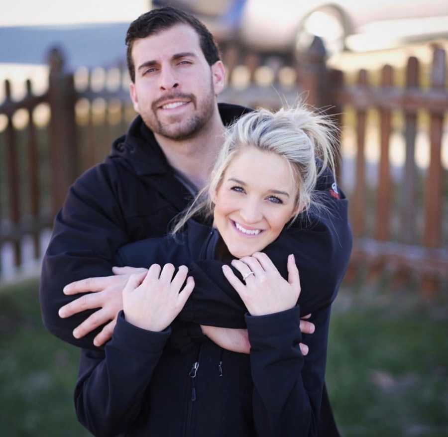 Husband stands with arms around wife who stands in front of him