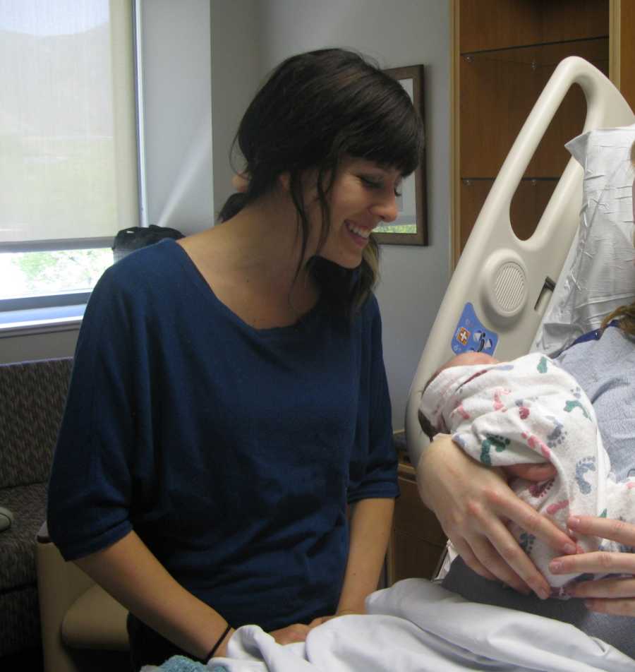 Woman stands beside hospital bed looking at newborn she is adopting in arms of birth mother