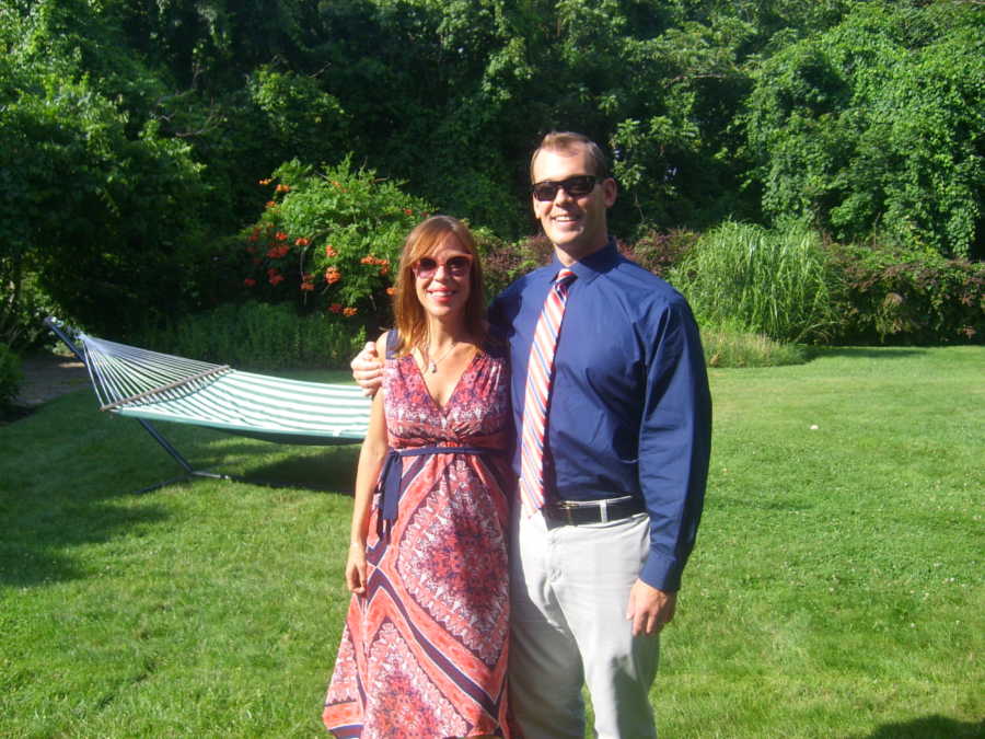 Husband stands in yard with arm around pregnant wife with hammock in background