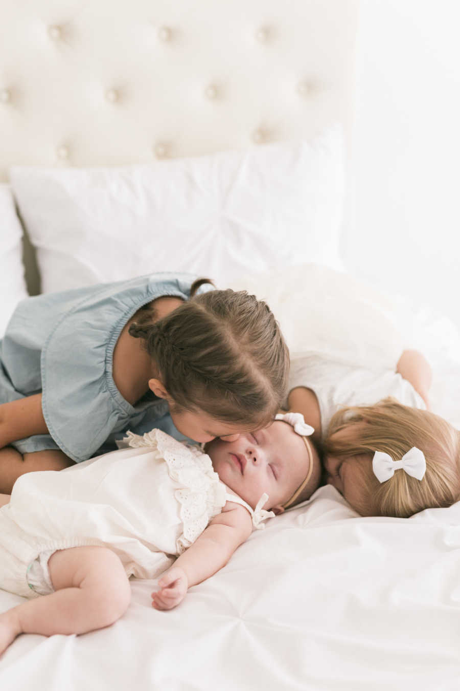 Newborn with down syndrome lays in bed while one older sister kisses her and other lays beside her