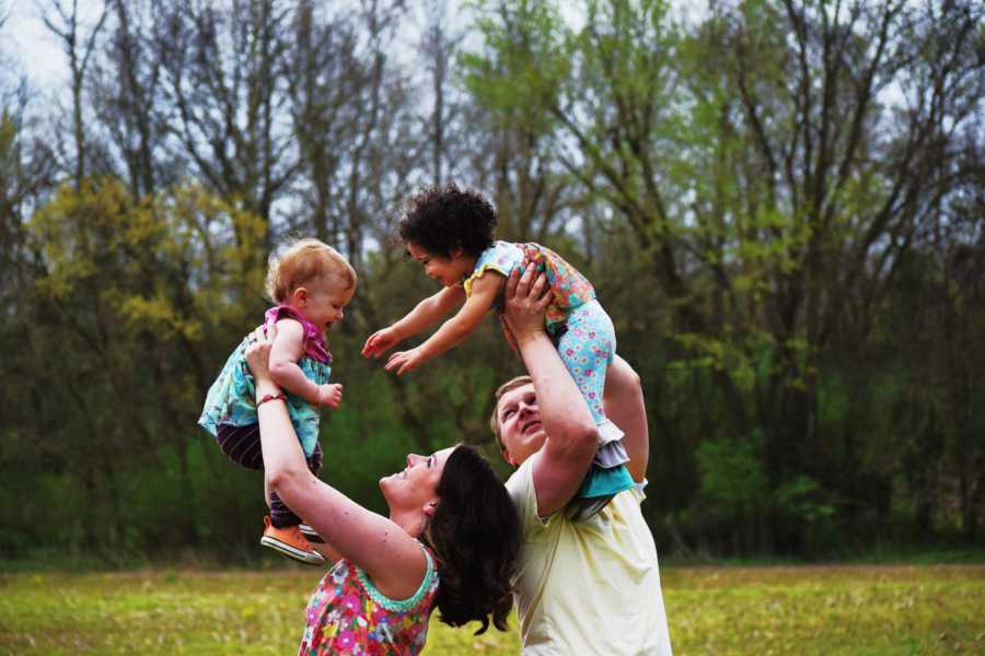 Husband and wife stand outside holding their adopted and biological daughter in air