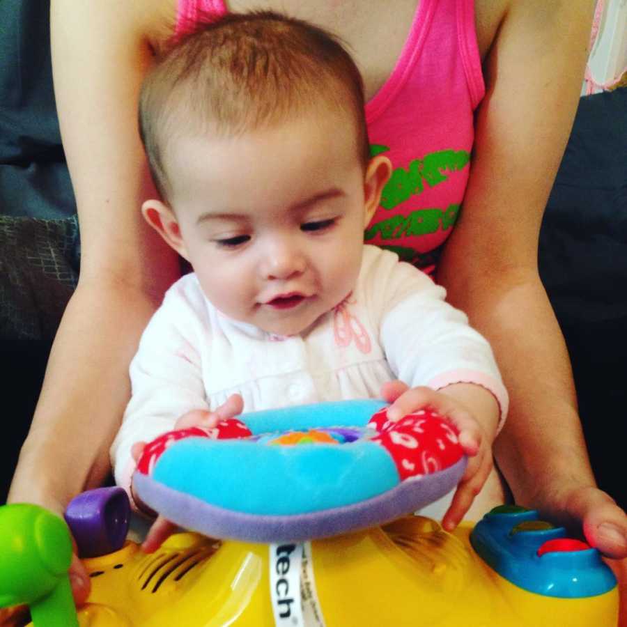 Baby sits playing with toy in lap of mother
