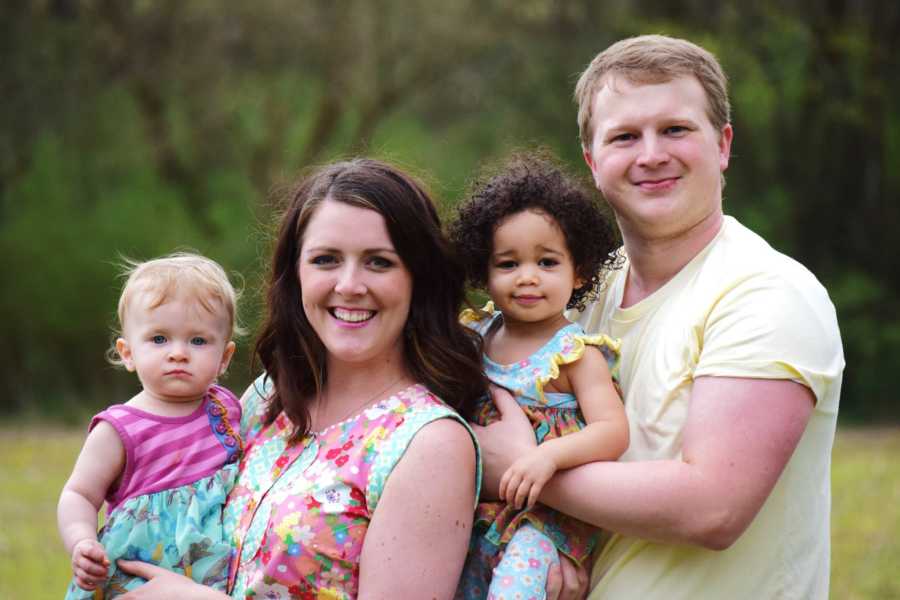 Husband and wife stand outside smiling while holding their adopted and biological daughters