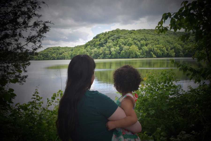 Mother holds adopted daughter as they stand in wooded area looking out at body of water