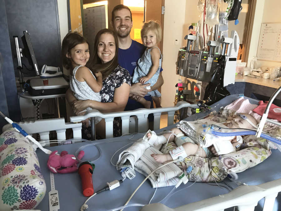 Baby with down syndrome and heart condition lays in PICU while her parents and two sisters stand over her