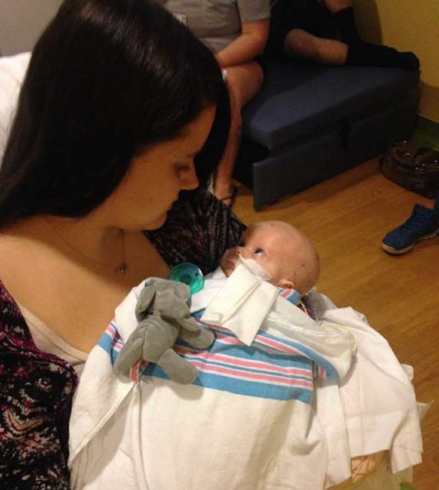 Mother sits in hospital holding newborn in her arms who has brain cancer