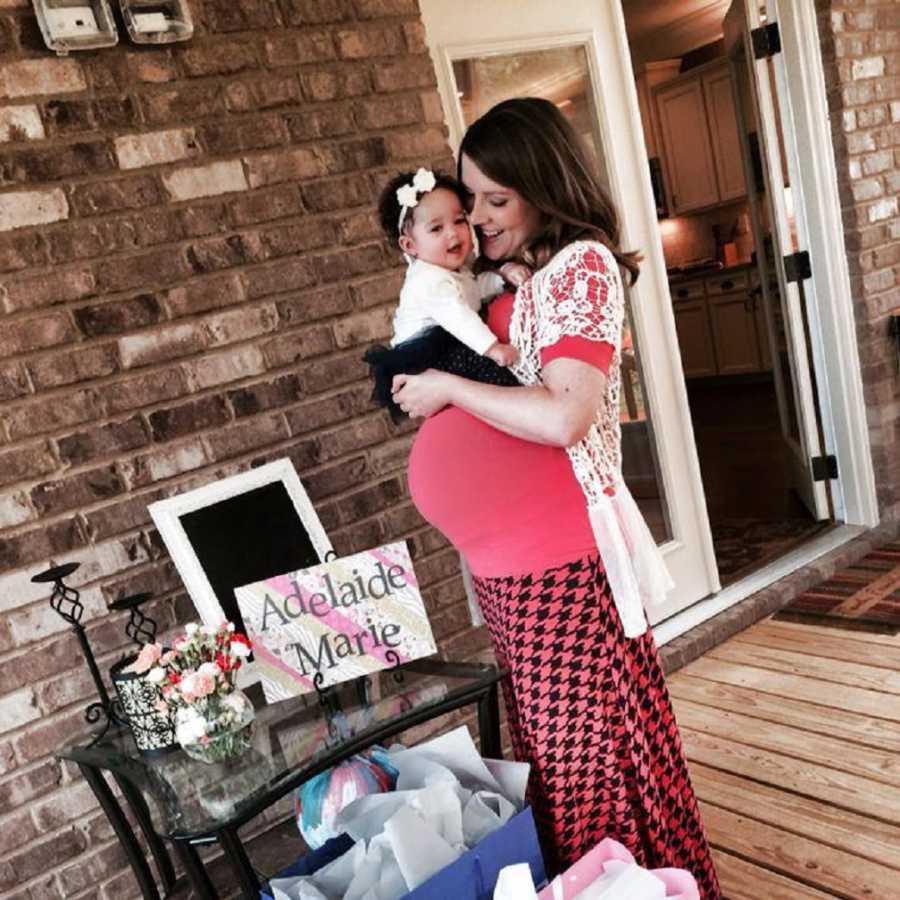 Pregnant woman stands outside home holding adopted baby girl