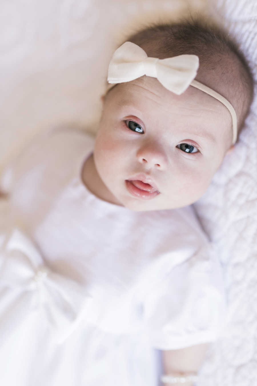 Baby with down syndrome and heart condition lays on back wearing white dress and headband