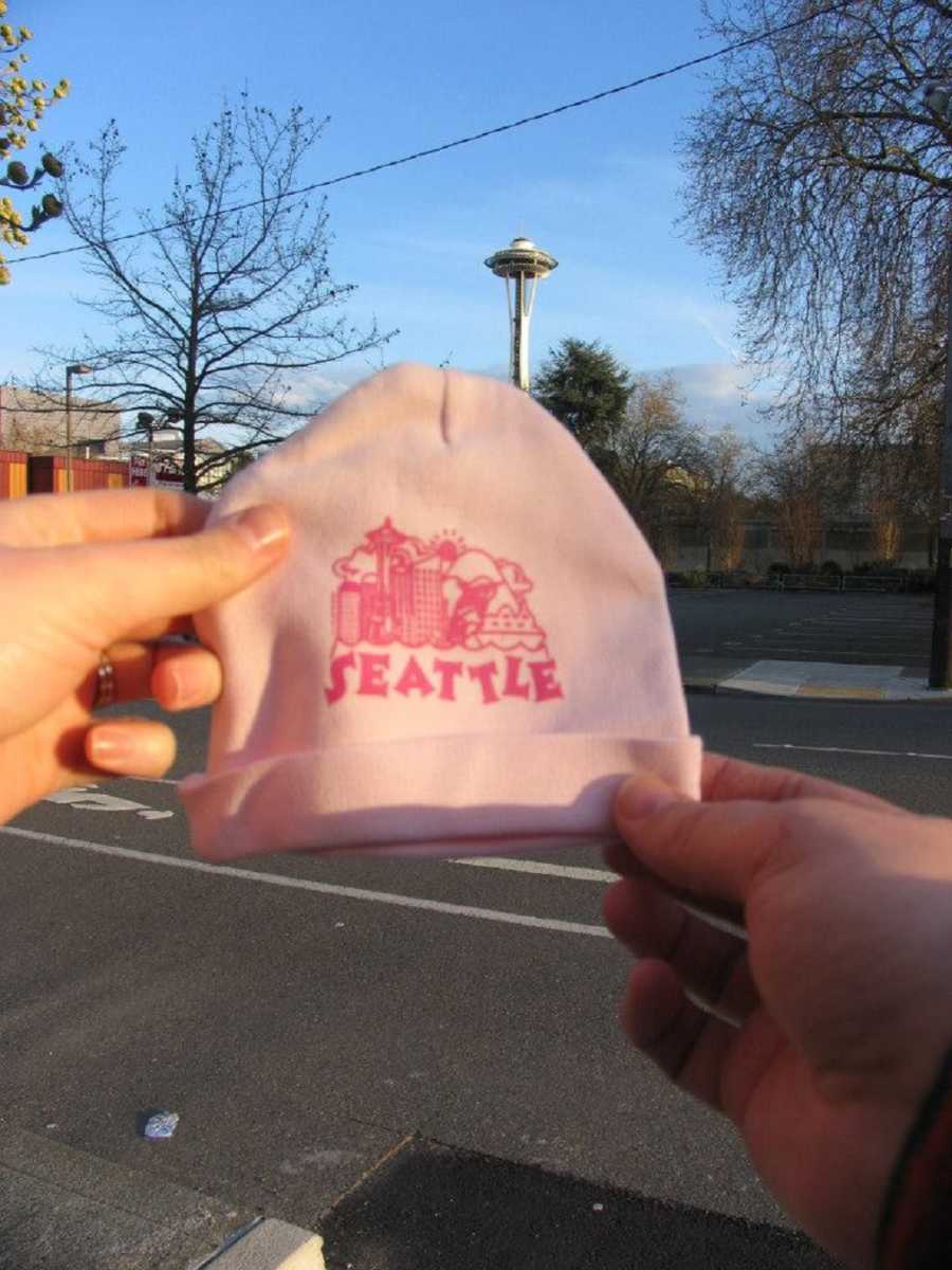 Husband and wife hold out pink baby hat that says," Seattle"