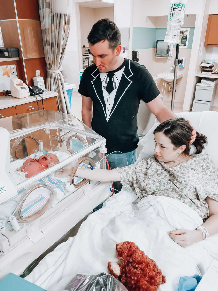 Mother sits reaching hand to crying newborn in NICU as father stands over them