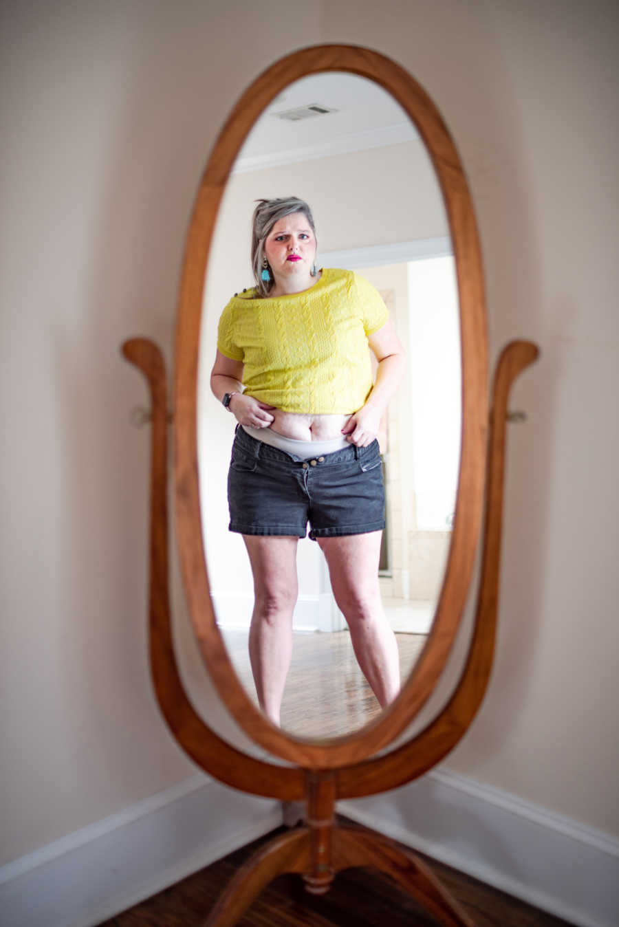 Mother who is hard on herself for her weight looks at herself in mirror with her stomach exposed